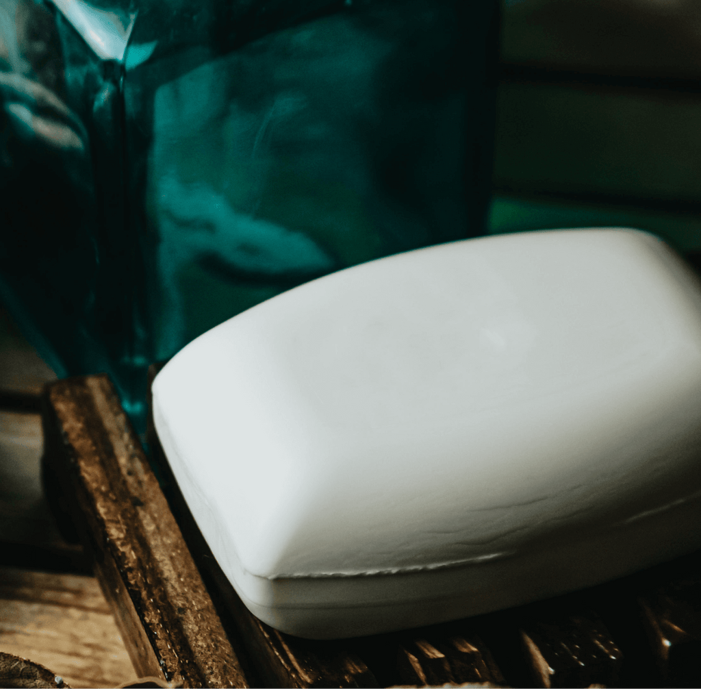 A white rectangular bar of soap resting on a dark wooden surface, with a blurred teal glass container in the background.